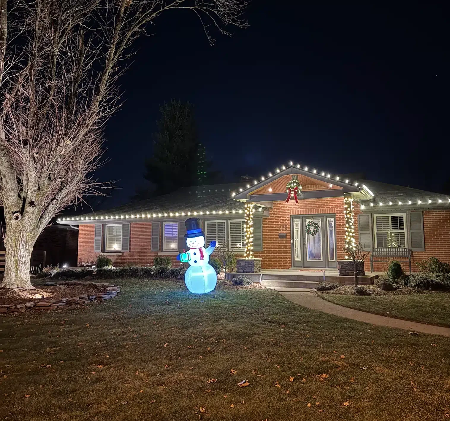 Brick ranch home with warm-white roofline lights, wrapped columns and a lit snowman at night