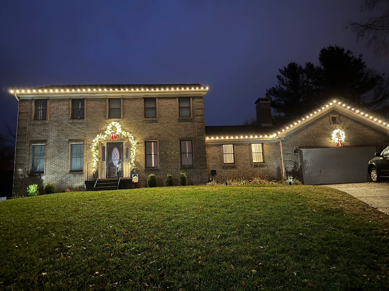 Brick colonial with warm-white roofline lights and door garland at dusk