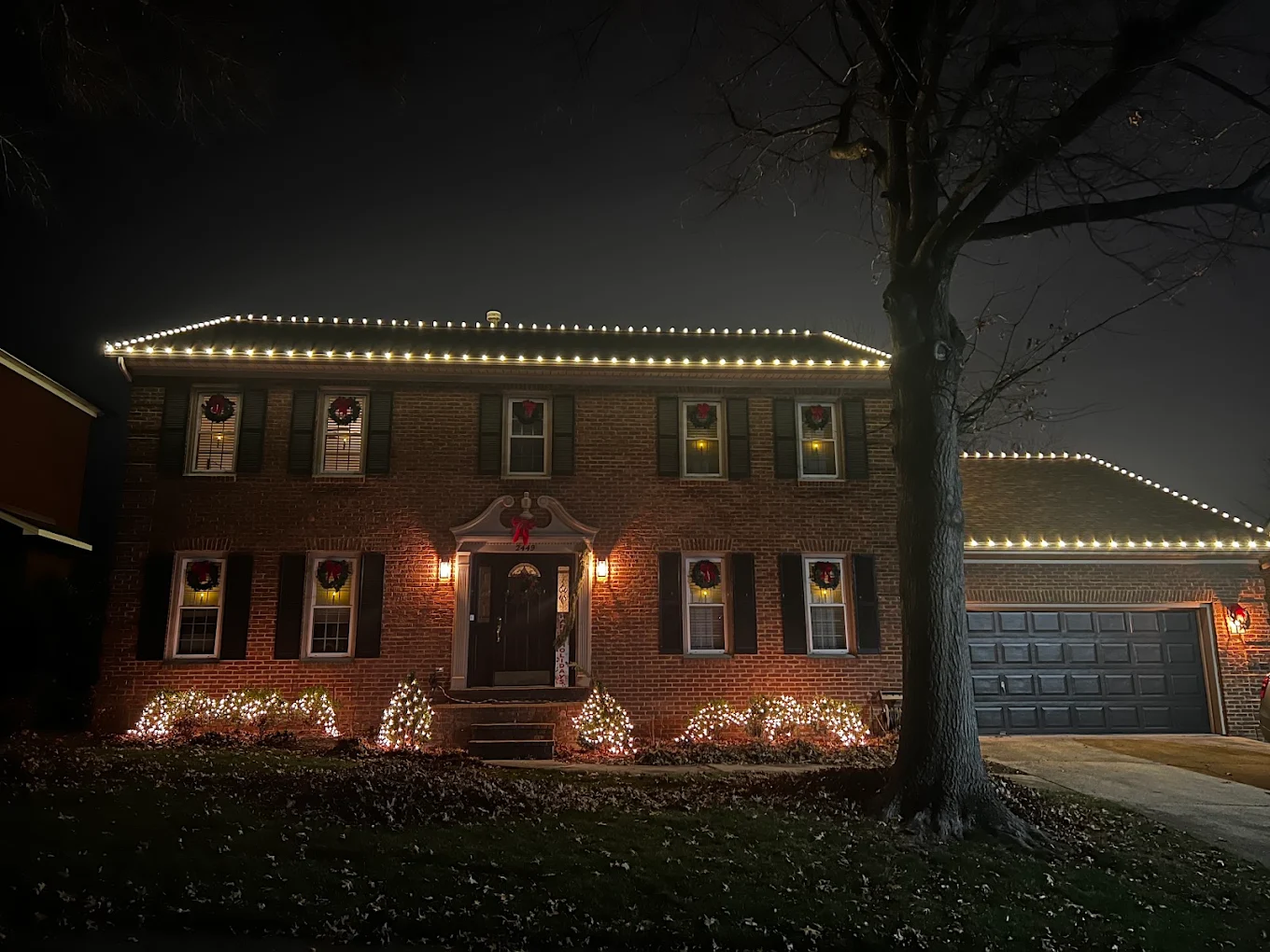 Brick colonial with roofline lights and lit wreaths in every window at night