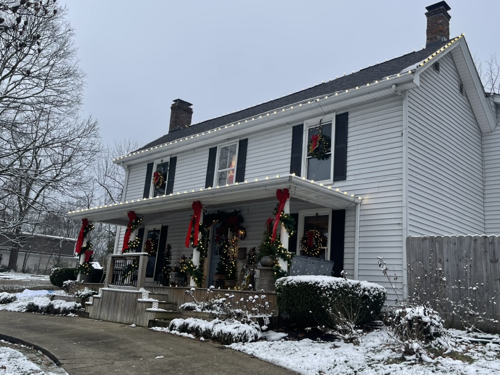 White farmhouse with roofline lights and red-bow garland in snow