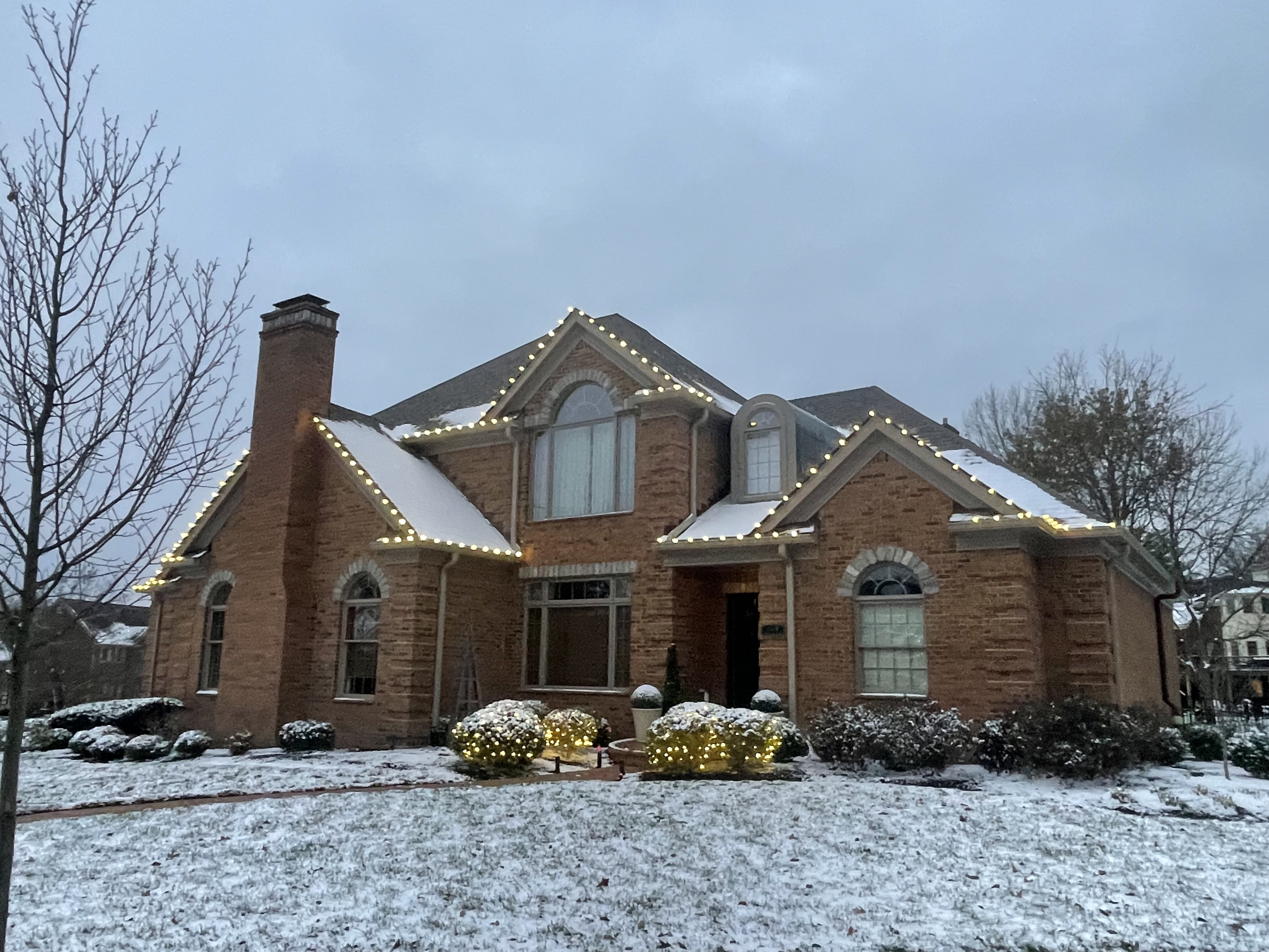 Brick home with roofline lights and lit shrubs in snow