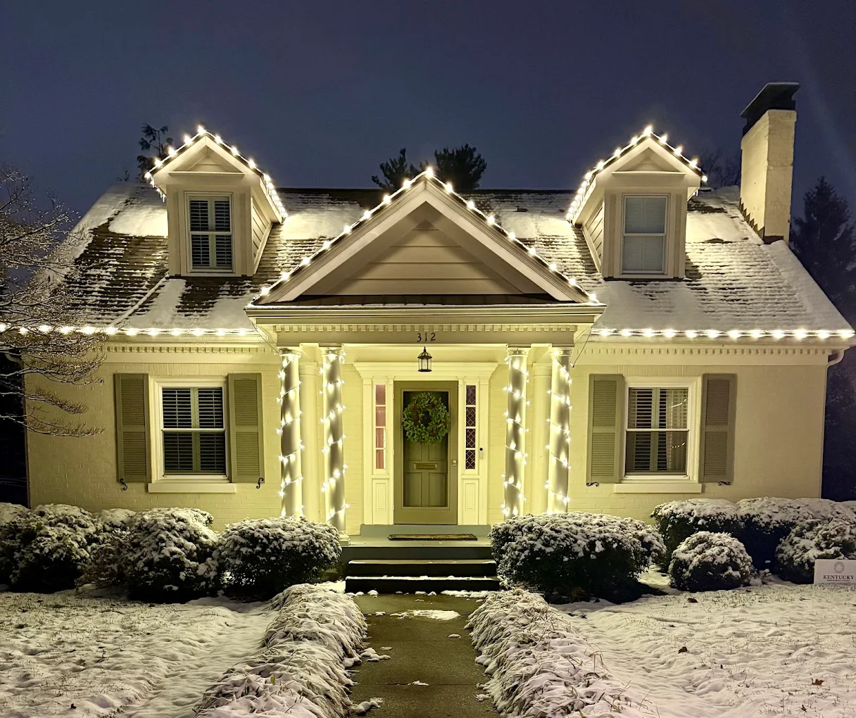 Cape Cod home with dormers, warm-white roofline and wrapped porch columns in snow at night