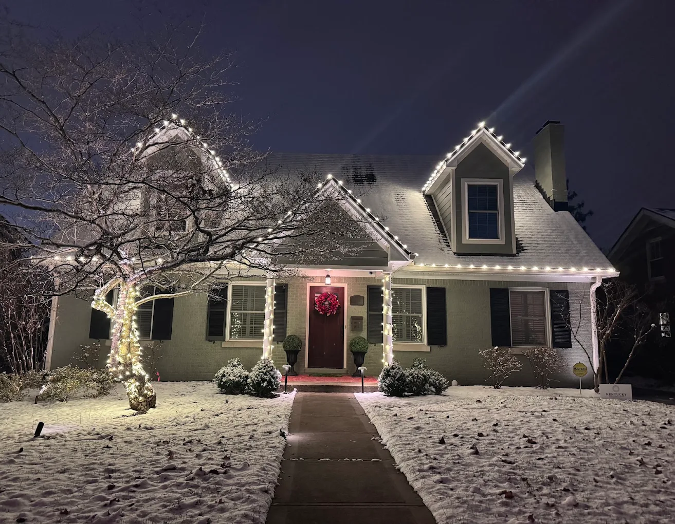 Cape Cod home with roofline lights, a wrapped tree and wrapped columns in snow at night