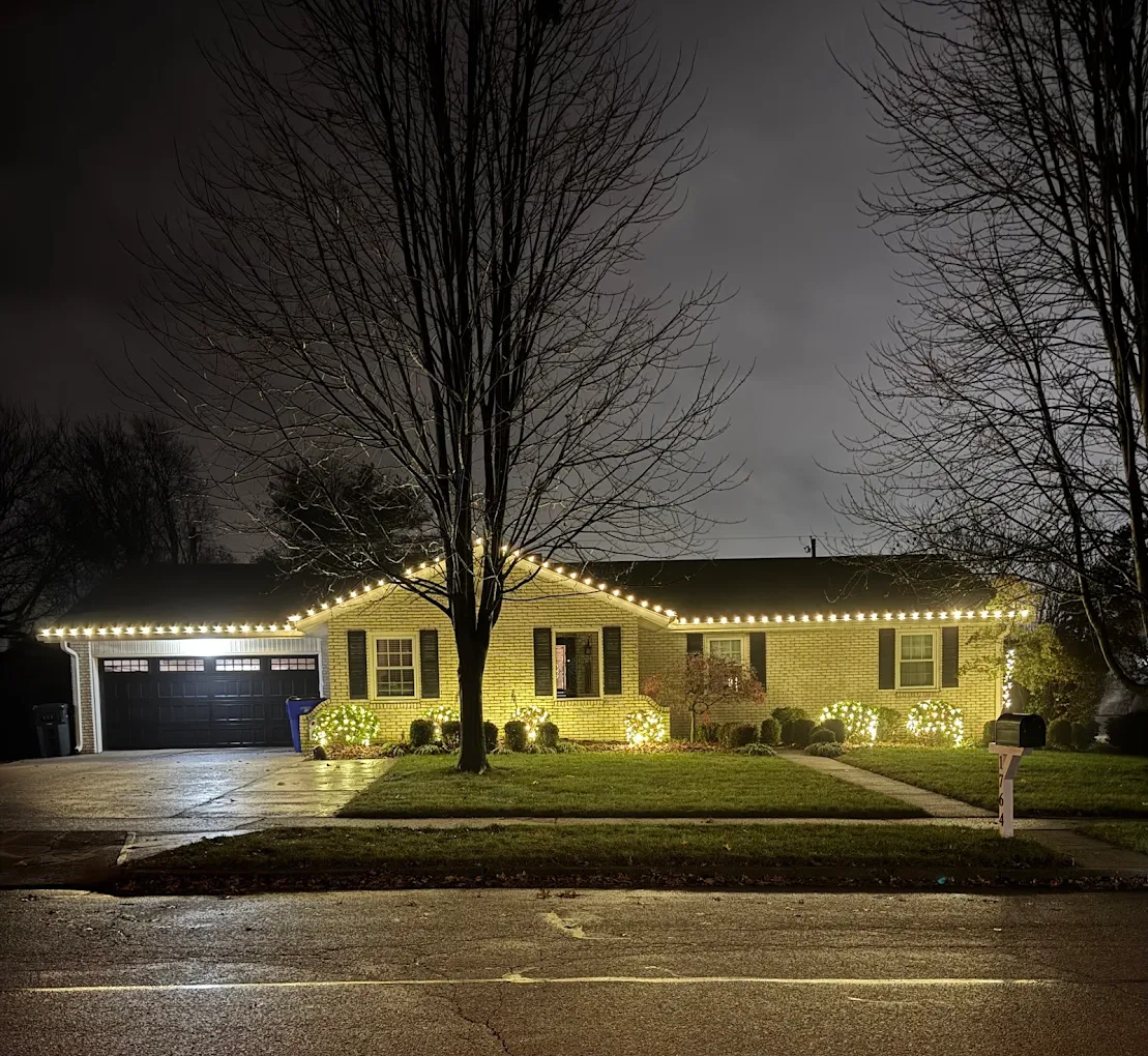 Brick ranch with warm-white roofline and lit shrubs at night